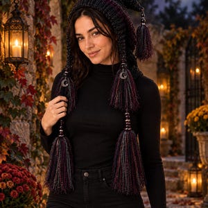 May include: A dark charcoal gray crocheted hood featuring a long pointed shape, adorned with two long hanging tassels in shades of black, purple, and gray. Each tassel is accented with silver-toned crescent moon charms and dark metallic beads.