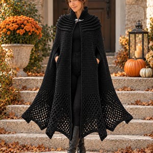 May include: A long, black, crocheted cape featuring a high collar with a silver-toned metal clasp closure. The garment has a solid upper section and a wide, open-work, lattice-style crochet pattern along the bottom hem, worn over black clothing.