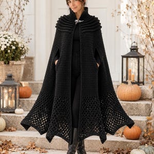 May include: A long, black, floor-length crochet cape featuring a high collar, a silver-toned metal clasp at the neckline, and a wide, intricate open-work lace pattern along the bottom hem. The garment has arm slits and a solid, dense stitch texture.