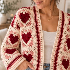 May include: A crochet cardigan featuring a repeating granny square pattern with dark red hearts centred on light pink and cream backgrounds. The garment has long sleeves, an open front design, and a ribbed cream border along the neckline and edges.