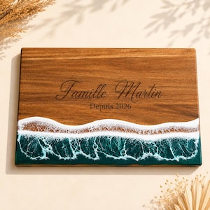 Peut inclure: Une planche à découper rectangulaire en bois avec un grain brun foncé. La partie inférieure est ornée d'un motif de vagues océaniques en résine turquoise et blanche. Le texte "Famille Martin" et "Depuis 2026" est gravé au centre.