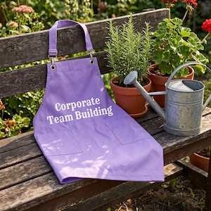 May include: A light purple apron featuring the white text Corporate Team Building printed on the front. The apron includes an adjustable neck strap with a metal buckle, two front pockets with metal rivets, and a straight hemline at the bottom.