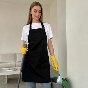 以下が含まれることがあります： 調節可能なバックル付きのネックストラップ、大きなフロントポケット、ストレートな裾が特徴の無地の黒いエプロン。白いTシャツと青いジーンズの上に着用し、鮮やかな黄色のゴム製クリーニンググローブを合わせています。