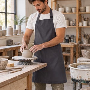 May include: A solid dark grey apron featuring a neck strap with an adjustable buckle and a large front pocket. The apron is constructed from a smooth, opaque fabric and provides full coverage from the chest down to the mid-thigh area.