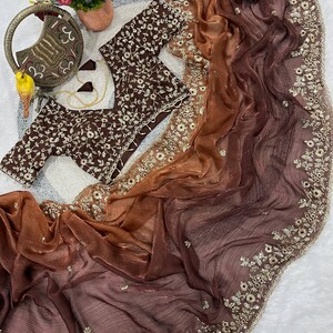Può includere: Una camicetta marrone scuro con intricati ricami floreali bianchi e un sari sfumato abbinato che passa dal rame al marrone scuro, caratterizzato da un ampio bordo con dettagliati ricami floreali dorati, paillettes e piccoli specchi.