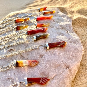 May include: Silver chain necklaces with pendants carved in the shape of a fist. The pendants are made of polished, banded agate in shades of red, orange, and brown. The necklaces are displayed on a gray stone surface.