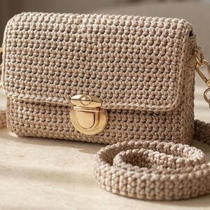 May include: A beige crochet crossbody bag featuring a rectangular flap, a gold-toned metal push-lock closure, and a matching thick crocheted shoulder strap attached with gold-colored metal lobster clasps on both sides of the bag.