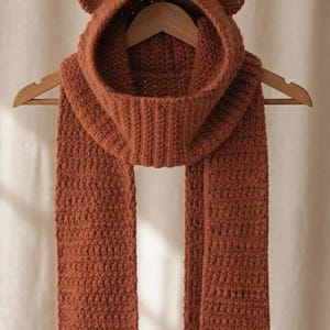 May include: A rust-coloured, chunky knit hooded scarf featuring two small, rounded bear ears on top of the hood. The scarf is constructed with a textured, ribbed stitch pattern and hangs vertically from a wooden hanger, displaying a uniform colour.
