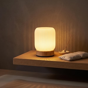 May include: A cylindrical table lamp with a ribbed, translucent white shade and a light-toned wooden base. The lamp emits a warm, soft glow, casting light patterns onto the surface. Next to it sits a small incense holder and a rectangular fabric pouch.