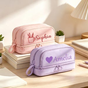 以下が含まれることがあります： Sophiaと書かれたペン立てのイラスト付きの薄いピンク色のペンケースと、Ameliaと書かれたハートのイラスト付きの薄い紫色のペンケース。どちらの長方形のケースも、2つのジッパー付きコンパートメントと円形のジッパープルを備えています。
