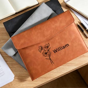 Op de afbeelding: Een bruine documentenmap met klepsluiting, voorzien van een zwarte lijntekening van een bloemenboeket en de naam William in het zwart gedrukt. De map ligt bovenop een grijze map en een zwarte map, alle met een gladde textuur.