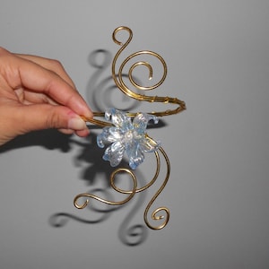 Puede incluir: Un brazalete con una estructura de alambre en tono dorado con intrincados patrones de espirales. Una flor de color azul claro translúcido e iridiscente con una pequeña cuenta blanca en el centro está unida a la parte frontal de la estructura.