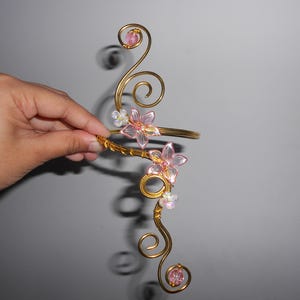 May include: A gold-toned wire arm cuff featuring intricate swirling metal patterns, accented with translucent pink flower-shaped beads and small white floral embellishments. The design incorporates circular wire loops at the top and bottom ends.
