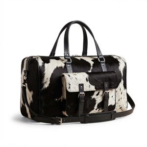 May include: A duffel bag featuring a black and white cowhide pattern with a textured hair-on-hide finish. The bag includes black leather handles, a detachable shoulder strap, and a front pocket secured by two buckled leather straps.