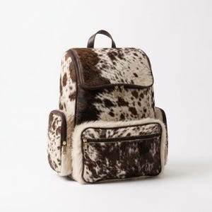 Puede incluir: Una mochila con un patrón de piel de vaca en marrón y blanco con un acabado de pelo texturizado. La bolsa incluye un compartimento principal grande con cremallera, un bolsillo frontal con cremallera y dos bolsillos laterales con ribetes marrones.