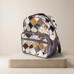 May include: A backpack featuring a patchwork design of cowhide squares in shades of brown, black, white, and tan. The bag includes a front zippered pocket, dark brown leather side panels, and silver-toned metal zippers with matching pull tabs.