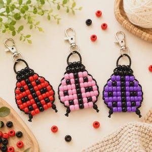 May include: Three ladybug-shaped keychains crafted from plastic pony beads on black cord, each featuring a silver-tone metal lobster clasp. The keychains are colored in red, light pink, and purple, all accented with black beads for spots.