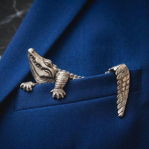 Può includere: Una spilla in metallo color argento a forma di coccodrillo fissata sulla tasca di un blazer blu intenso. La figura del coccodrillo presenta squame dettagliate, una testa testurizzata e una lunga coda che si estende sull'apertura della tasca.