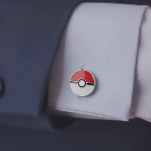 May include: A circular cufflink featuring a red and white design with a black horizontal stripe and a central black circle, resembling a Poké Ball. The cufflink is fastened through the buttonhole of a light blue shirt cuff, partially covered by a dark blue jacket sleeve.