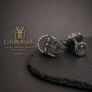 Puede incluir: Un par de gemelos de metal plateado con el diseño de la nave espacial Millennium Falcon en un lado y el símbolo de la Alianza Rebelde en el otro. El texto GiftByFinds Cufflinks Brooches Necklaces Rings Earrings es visible.