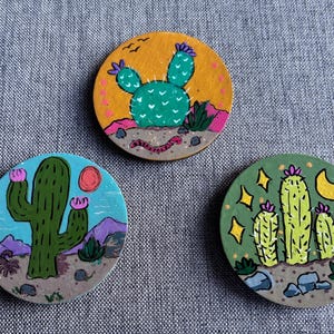 May include: Three circular wooden coasters featuring hand-painted desert scenes. One shows a green cactus on a yellow background, another a green cactus under a red sun, and the third three green cacti under a yellow crescent moon and stars.