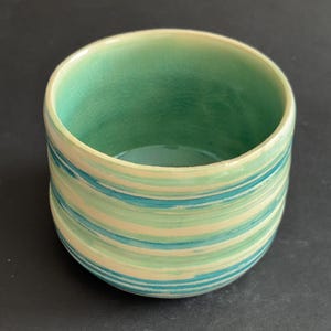 May include: A ceramic cup featuring a glossy finish with a crackle glaze texture. The exterior displays horizontal stripes in shades of light green, cream, and teal. The interior is a solid, smooth, light green colour with a visible crackle pattern.
