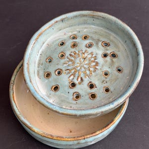May include: A two-piece ceramic soap dish featuring a light blue glazed top tray with a central snowflake-like embossed pattern and multiple drainage holes. The bottom base is a smooth, solid light beige bowl with a matching light blue rim.