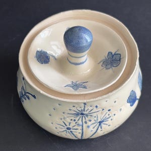 May include: A round, off-white ceramic lidded jar featuring a blue floral pattern. The lid has a central blue knob handle and small blue flower motifs. The base of the jar displays larger blue dandelion-like botanical illustrations and textured dots.
