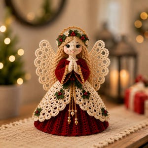 May include: A crocheted angel figurine with long blonde hair, wearing a deep red dress with a cream-colored lace overlay. The angel features intricate lace wings, a halo adorned with holly berries and leaves, and hands clasped in prayer.