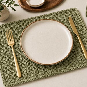 May include: A rectangular sage green crocheted placemat featuring a textured stitch pattern. Centered on the placemat is a round, speckled off-white ceramic plate with a brown rim, flanked by a matte gold fork on the left and a gold knife on the right.
