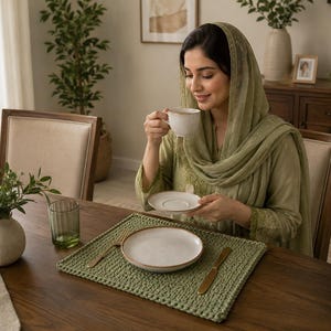 May include: A sage green crocheted rectangular placemat featuring a textured knit pattern, displayed on a wooden table with a white ceramic plate, a gold fork, a gold knife, a green glass tumbler, and a small white ceramic vase with green leaves.