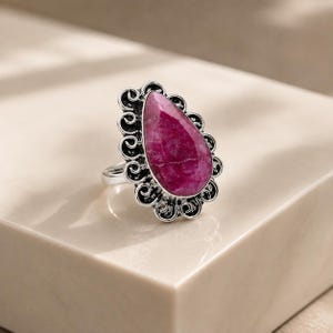 May include: A silver ring featuring a large, pear-shaped, deep pink gemstone set in a decorative metal frame with intricate swirling patterns. The band is plain silver, and the setting has a dark, oxidized finish to highlight the metal details.