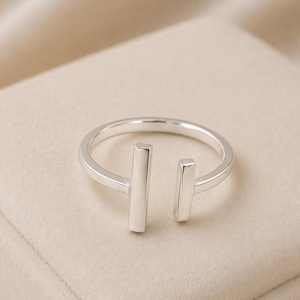 May include: A silver-toned open ring featuring two vertical rectangular bars at the ends. The band is smooth and polished, with a gap between the two parallel bars. The metal has a reflective surface, creating a clean and simple design.