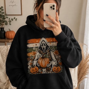 May include: A black hooded sweatshirt featuring a graphic print of a cloaked figure with a white mask holding a pumpkin, surrounded by autumn leaves, flowers, and two additional jack-o'-lanterns against a background of horizontal orange stripes.
