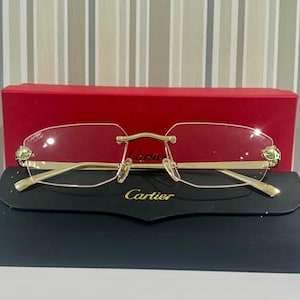 May include: Rimless Cartier spectacles featuring gold-tone metal temples and bridge, with panther head accents on the outer edges of the clear lenses. The glasses are displayed in front of a red Cartier box and a black Cartier case with gold text.