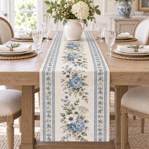 Puede incluir: Un camino de mesa rectangular con un patrón central vertical de flores de hortensia azules y anémonas blancas con follaje verde, flanqueado por bordes de rayas azules y blancas sobre una base de tela de color beige claro, centrado en una mesa de madera.