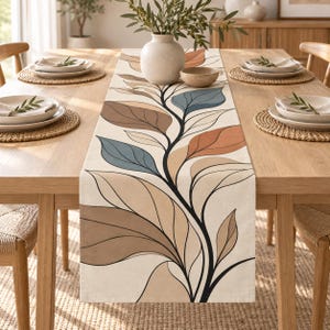 May include: A rectangular table runner featuring a cream-coloured fabric base with a central botanical print. The design displays stylised leaves in shades of terracotta, muted blue, and various tones of brown, all outlined in bold black lines.