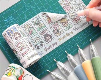Puede incluir: Un rollo de cinta washi blanca con una cuadrícula de ilustraciones de dibujos animados, números y el texto "Happy Monday Tuesday". Un segundo rollo muestra formas geométricas e iconos de comida. Cinco bolígrafos de gel con agarres pastel.