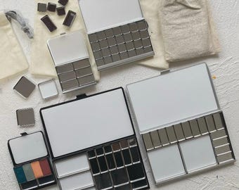 Puede incluir: Una colección de cajas metálicas para acuarelas de varios tamaños, algunas abiertas que muestran áreas de mezcla blancas vacías y filas de godets rectangulares, con varios godets individuales sueltos esparcidos y bolsas de tela beige con cordón.