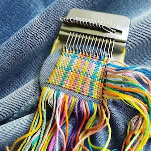 Peut inclure: Un outil de métier à repriser en métal avec une base rectangulaire et une rangée de crochets sur le dessus, retenant un carré de tissu tissé. Le tissage présente un motif multicolore vif composé de fils jaunes, orange, bleus et roses.