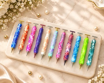 Puede incluir: Una colección de once bolígrafos con cuerpos de acrílico jaspeado multicolor y tapas metálicas en forma de T. Los bolígrafos presentan varias combinaciones de colores, incluyendo azul, naranja, rosa, morado, melocotón, azul claro, magenta, rojo y verde.