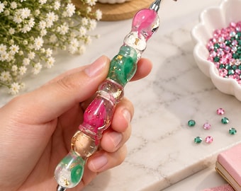 Puede incluir: Un bolígrafo para pintura de diamantes con mango de resina transparente y secciones alternas en rosa y verde, incrustado con copos de pan de oro. La herramienta tiene una punta de metal plateado y un accesorio metálico plano en forma de T.