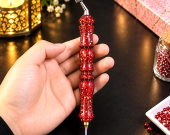 Puede incluir: Un bolígrafo para pintura de diamantes con un cuerpo de resina roja vibrante lleno de purpurina brillante. La herramienta tiene un mango contorneado de varias secciones y una punta metálica de color iridiscente, con un pequeño accesorio de metal en la parte superior.