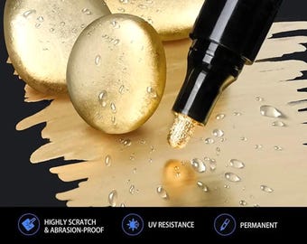 Puede incluir: Un marcador de pintura negro y dorado con punta metálica, junto a dos piedras doradas lisas cubiertas de gotas de agua. El marcador incluye el texto HIGHLY SCRATCH & ABRASION-PROOF, UV RESISTANCE, PERMANENT, HIGH-GLOSS y HIGHLY PIGMENTED SPECIAL INK.
