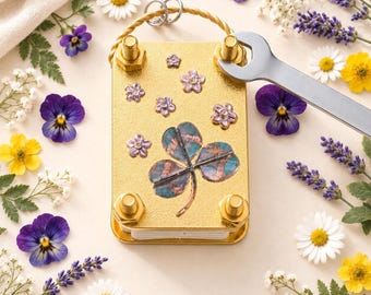 Puede incluir: Una prensa de flores rectangular de metal dorado con un diseño central de trébol de tres hojas y pequeños detalles florales. La prensa está asegurada con cuatro pernos en las esquinas y una llave plateada está colocada en el perno superior derecho.