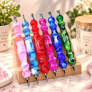 Può includere: Sei penne per diamond painting con impugnature in acrilico sagomate e multicolori in rosa, azzurro, rosso, magenta, blu scuro e verde. Ogni penna presenta una punta metallica e una clip in metallo argentato, esposte su un supporto in legno.