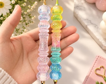 Puede incluir: Dos bolígrafos para pintura de diamantes con cuerpos translúcidos, texturizados y brillantes. Uno es rosa claro con punta y parte superior de metal dorado, el otro tiene un degradado arcoíris de amarillo, rosa, verde y azul con piezas metálicas iridiscentes.