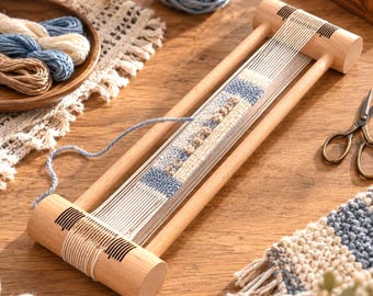 Puede incluir: Un telar rectangular de madera con dos extremos cilíndricos que presentan marcas negras. El telar sostiene hilos de urdimbre blancos y una tira textil parcialmente tejida en tonos azul, crema y beige, con un hilo de lana azul suelto.