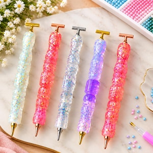 May include: Five diamond painting pens with textured, glitter-filled resin barrels in white, pink, clear, purple, and coral. Each pen features a metallic T-shaped top and a pointed metal tip, designed for placing small craft beads onto a surface.