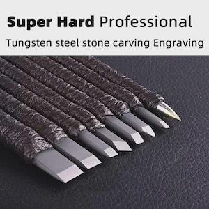 May include: A set of tungsten steel stone carving and engraving tools with dark, textured wrapped handles. The tools feature various metal tip shapes, including flat, angled, and pointed ends, arranged in a row for detailed stone work.